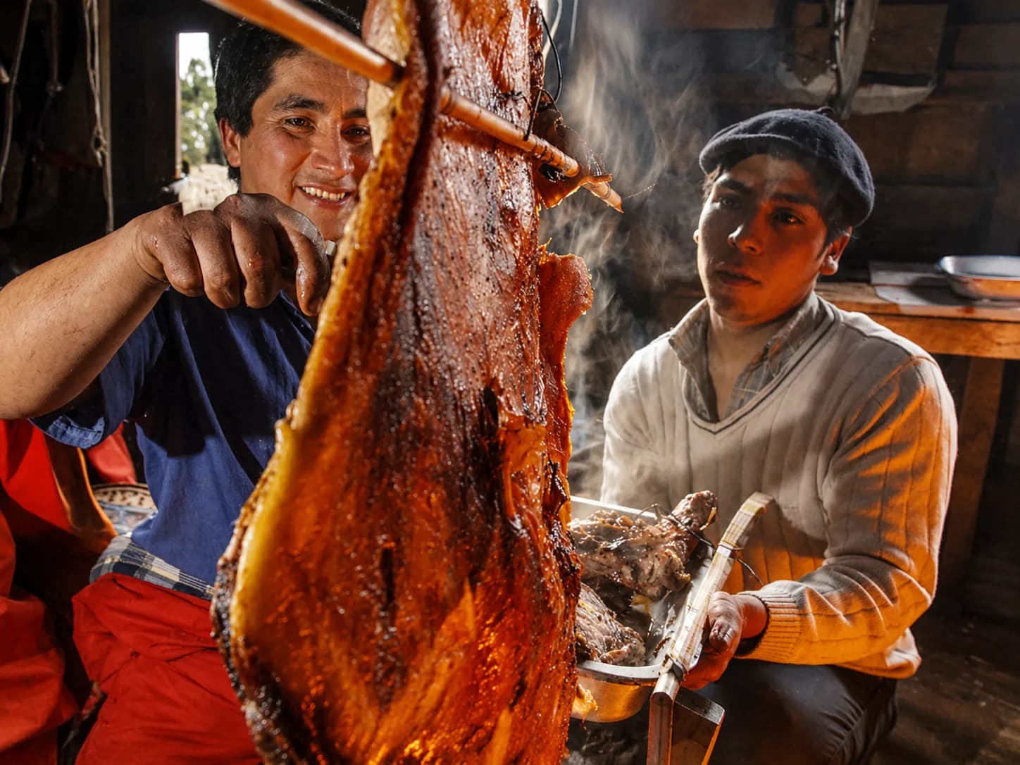 Dos cocineros sonriendo junto a un cordero asado al palo