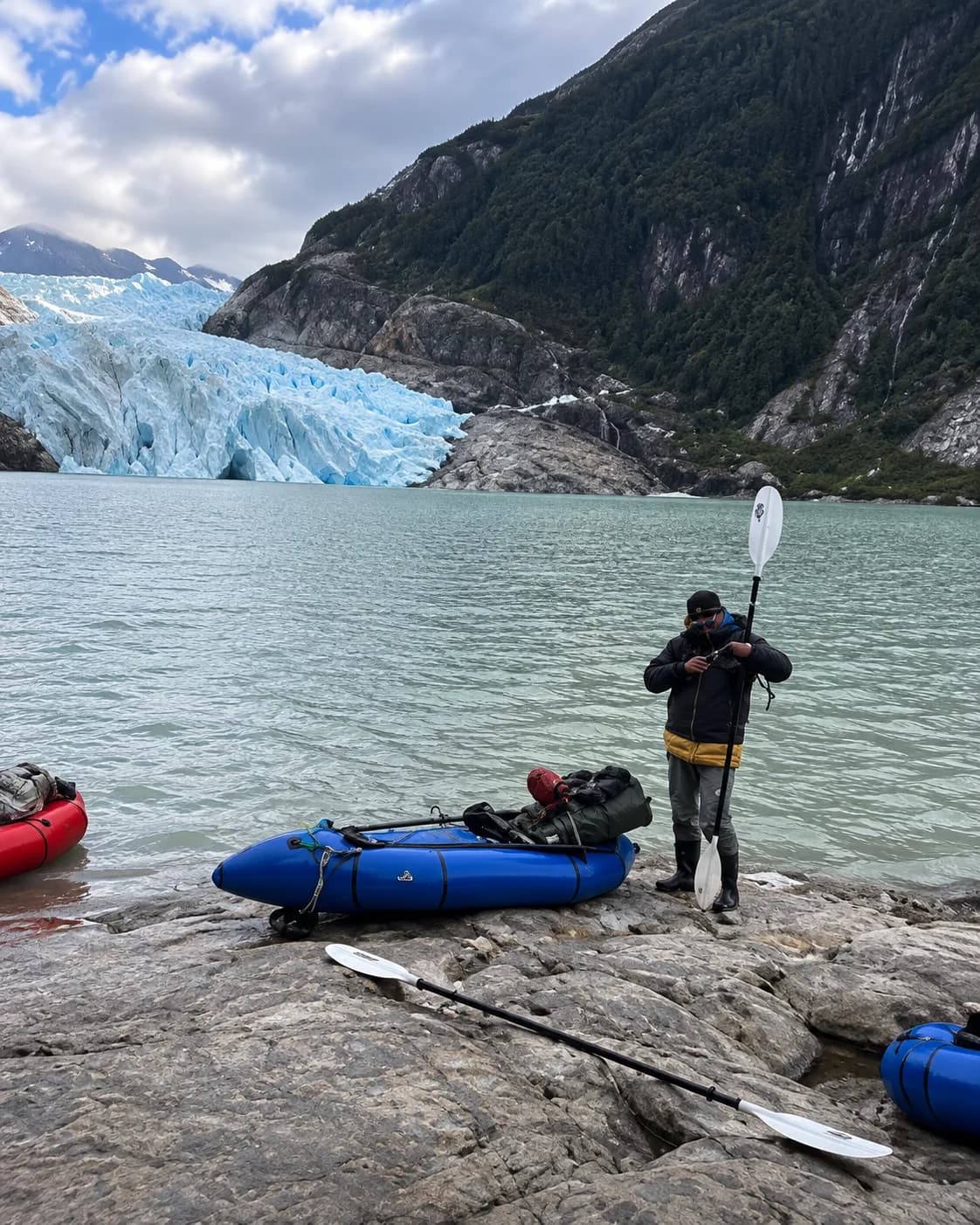 Persona junto a un packraft azul en la orilla rocosa de un fiordo, con un glaciar al fondo