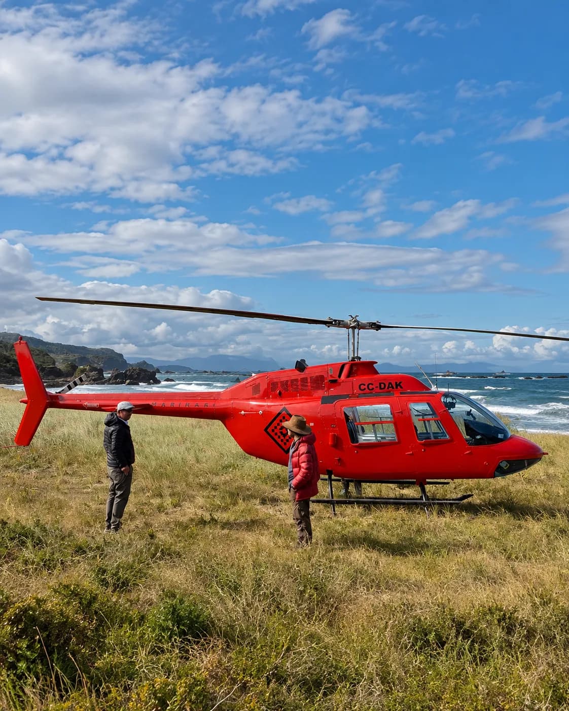 Helicóptero rojo posado en un pastizal junto a la costa, con dos personas al lado