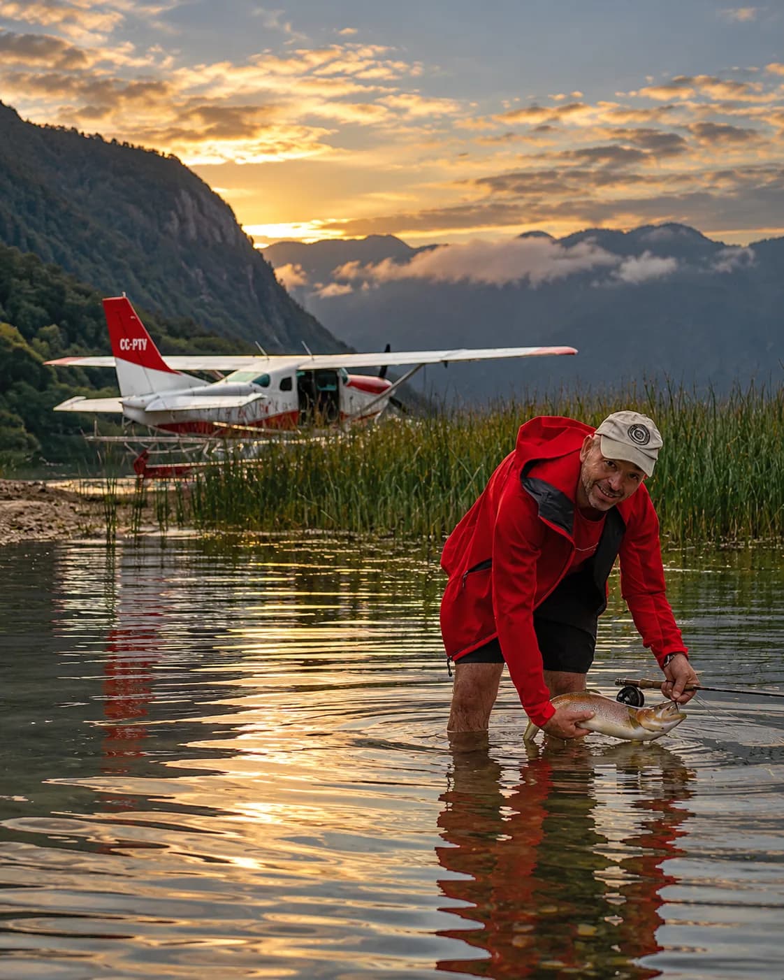 Pescador con mosca sosteniendo una trucha en aguas poco profundas, con un hidroavión al fondo al atardecer