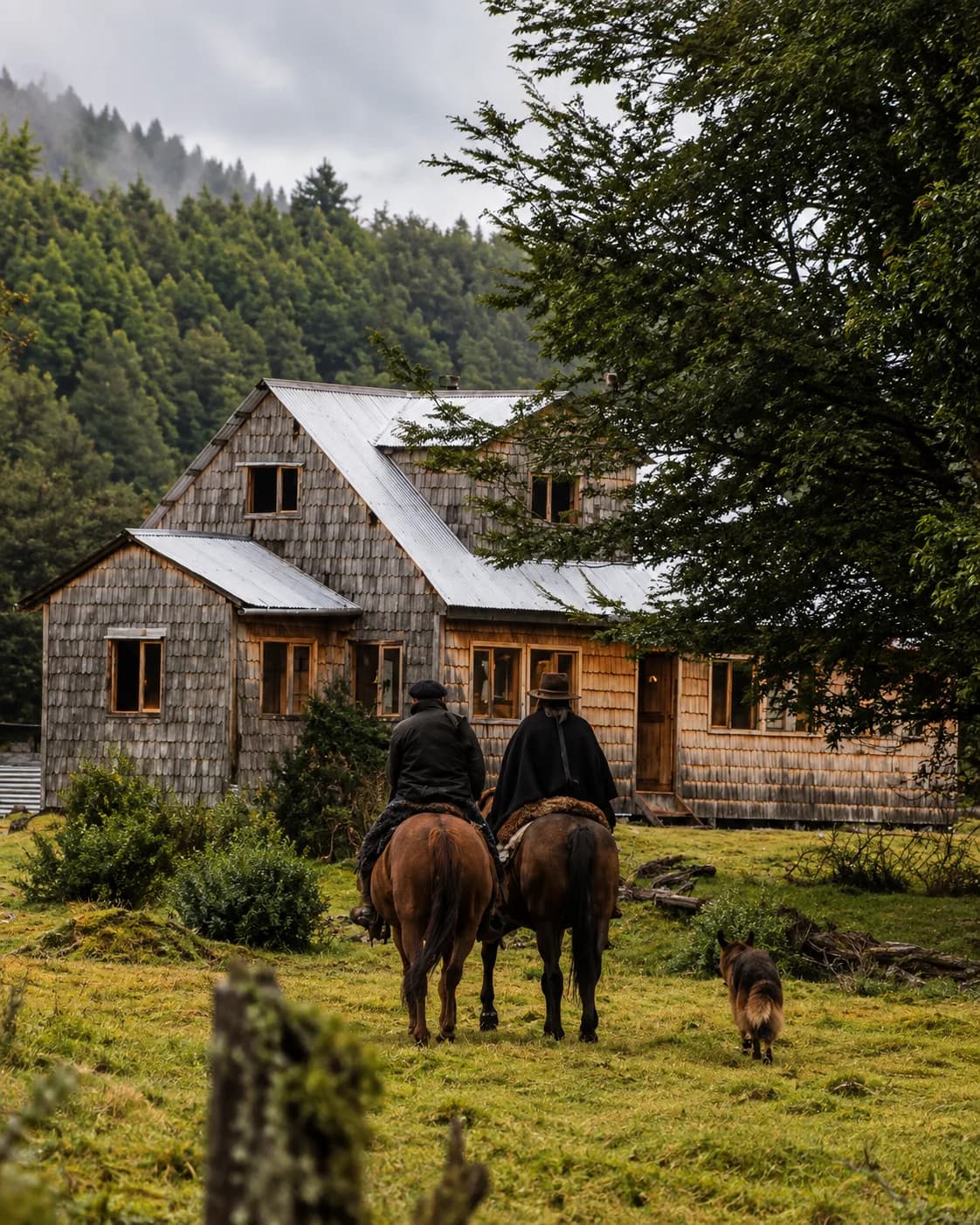Dos jinetes a caballo frente a una casa de madera con techo de zinc, en medio del bosque