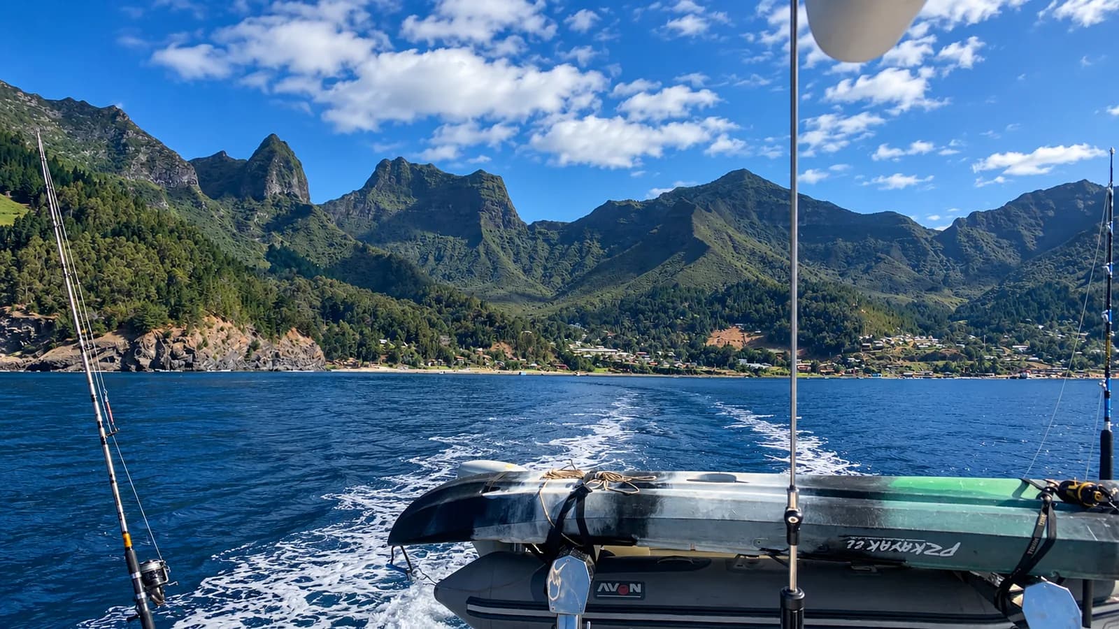 Bote inflable de apoyo con cañas de pesca, dejando estela frente al poblado de San Juan Bautista entre montañas volcánicas